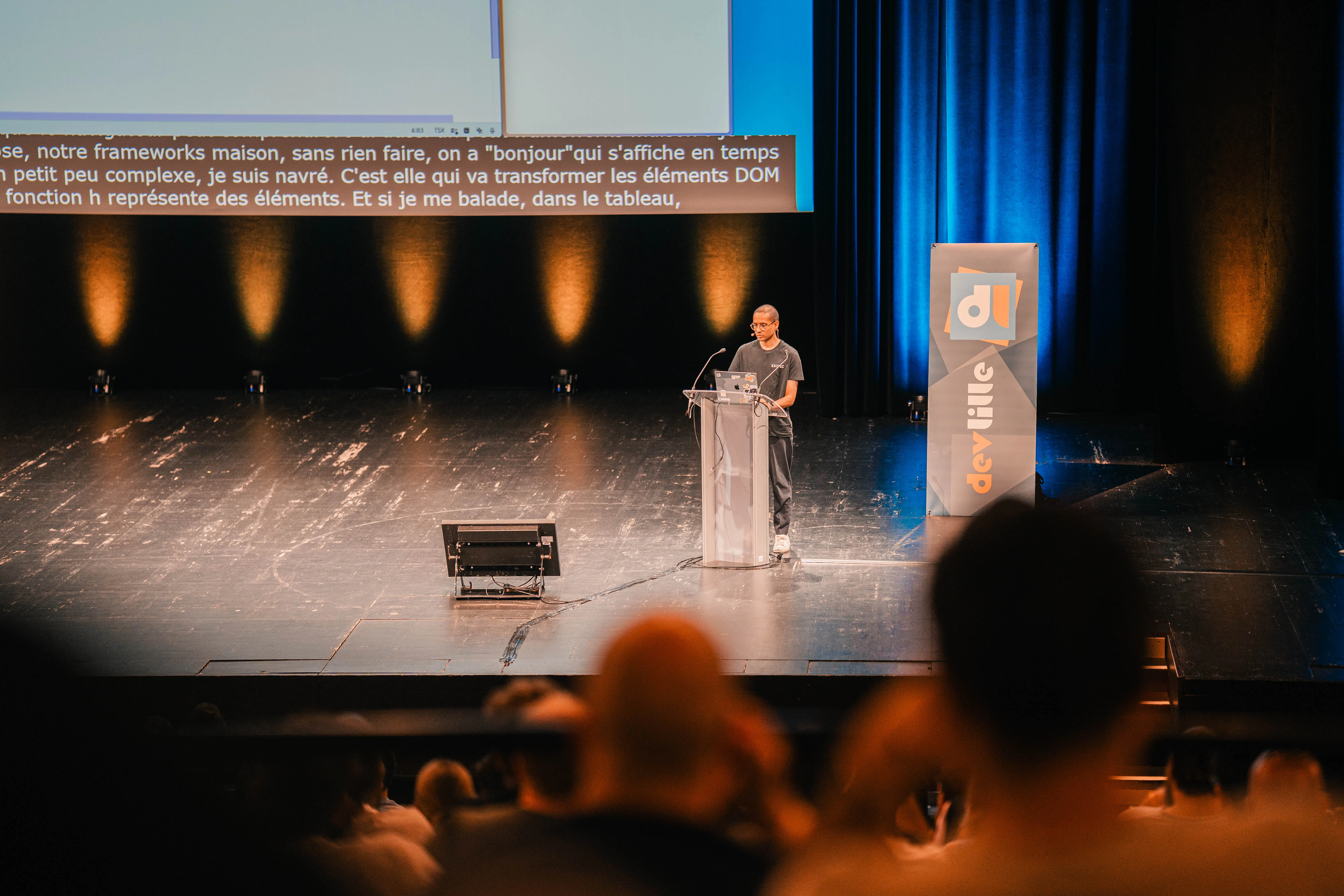 je suis sur la scène d'un amphithéâtre dans une salle de conférence, je parle derrière un publique. au premier plan, des personnes floues assises sur les gradins qu'on voit de dos